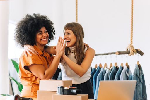 Two female business owners giving each other a high five while smiling with boxes in front of them and clothes hanging in the background.
